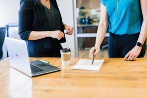 Two women stand at a wooden table with a laptop, coffee cup, and notepad; one points at the notepad with a pen while the other listens, suggesting a discussion or meeting in an office setting.