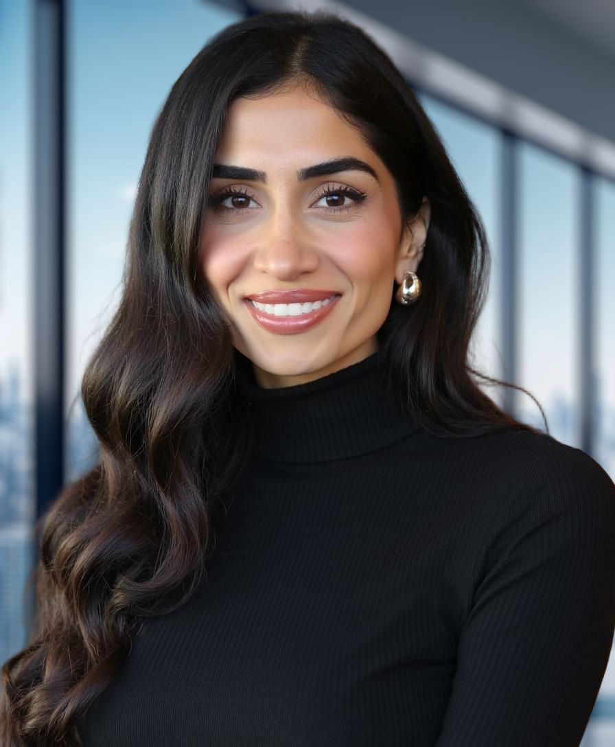 A woman with long, wavy dark hair and hoop earrings smiles while wearing a black turtleneck. She stands in front of large windows with a cityscape in the background.